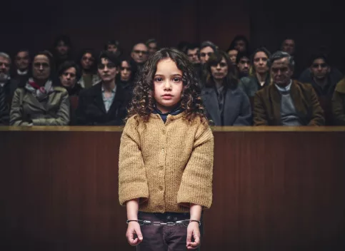 Une petite fille est debout, menottes aux poignets. Derrière elle on aperçoit des personnes assises dans un tribunal